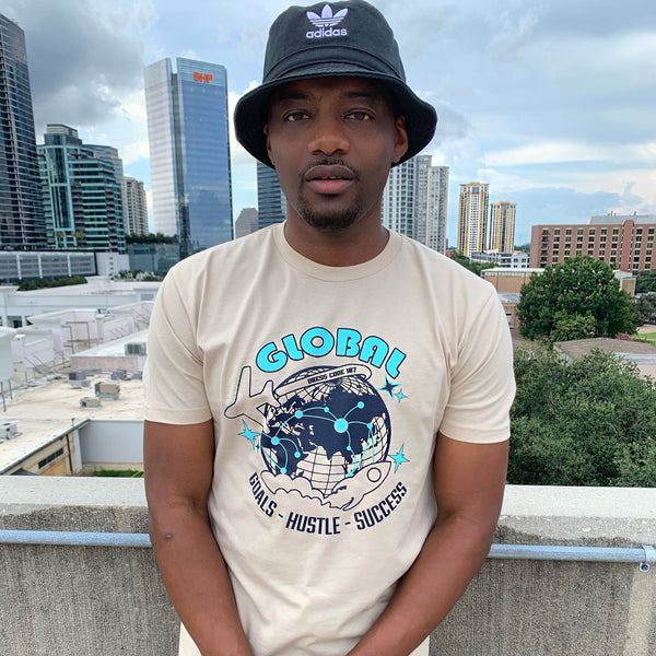 Cream base t-shirt. The design is navy blue and teal blue. The words "global" appear in bubble bold letters. The graphic is a globe, air plane and rocket. The text goals hustle and success appear under the globe. 